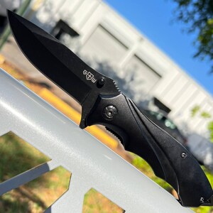 May include: A black folding knife with a sharp blade. The knife has a textured handle and a small emblem near the blade's base. The knife is shown against a blurred background of a building and trees.