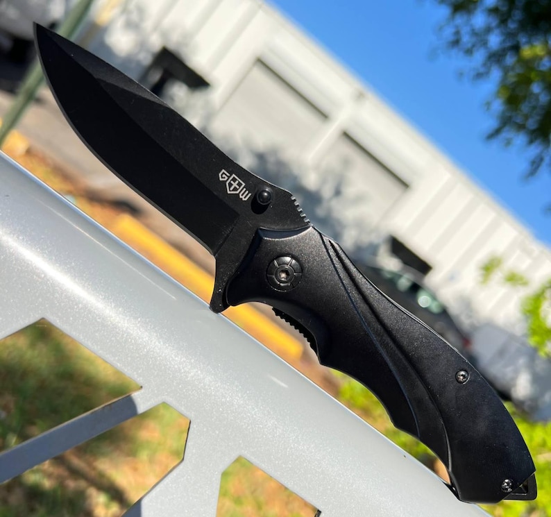 May include: A black folding knife with a sharp blade. The knife has a textured handle and a small emblem near the blade's base. The knife is shown against a blurred background of a building and trees.