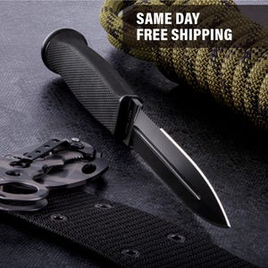 May include: A black tactical knife with a textured handle and a partially serrated blade. The knife is displayed with a black belt and a camouflage multi-tool. A green and brown rope is in the background.