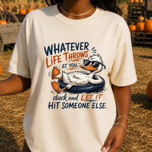May include: A cream-colored t-shirt featuring a graphic of a white duck wearing black sunglasses while floating in a dark blue inner tube. The text reads: WHATEVER LIFE THROWS AT YOU, duck and LET IT HIT SOMEONE ELSE. The design uses blue and orange ink.