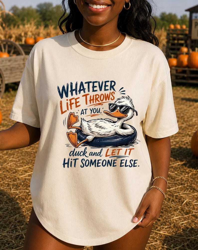 May include: A cream-colored t-shirt featuring a graphic of a white duck wearing black sunglasses while floating in a dark blue inner tube. The text reads: WHATEVER LIFE THROWS AT YOU, duck and LET IT HIT SOMEONE ELSE. The design uses blue and orange ink.