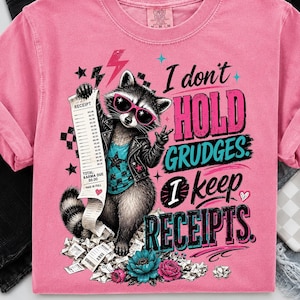 May include: A pink t-shirt featuring a graphic of a raccoon wearing a black leather jacket and pink sunglasses. The raccoon holds a long receipt. The text reads: I don't HOLD GRUDGES. I keep RECEIPTS. The design includes stars, lightning, and flowers.