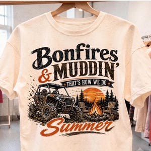 May include: A cream-coloured short-sleeve t-shirt featuring a graphic print of an off-road vehicle, a campfire, and pine trees. The text reads Bonfires & MUDDIN' THAT'S HOW WE DO Summer in black and orange lettering with a sunset background.
