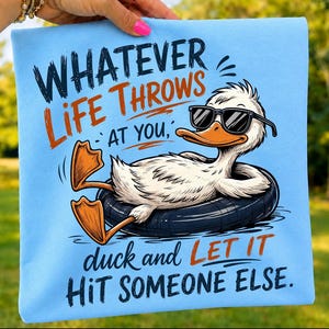 Folded light blue t-shirt mockup showcasing a funny duck wearing black sunglasses relaxing in an inner tube with the quote "Whatever Life Throws At You, Duck and Let It Hit Someone Else," in bold retro typography