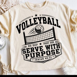 Op de afbeelding: Een crèmekleurig T-shirt met een zwarte afbeelding van een volleybal, een net en de tekst "VOLLEYBALL", "DIG DEEP", "TRUST GOD", "SERVE WITH PURPOSE" en "ISAIAH 40:31". Het ontwerp bevat sterren en lijnen op de voorzijde van het shirt.