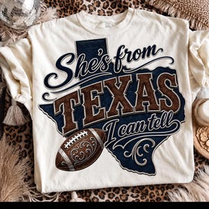 May include: A cream-colored t-shirt featuring a dark blue graphic of the state of Texas. The text reads "She's from TEXAS I can tell" in dark blue and brown lettering. A brown football with silver filigree accents is positioned at the bottom.