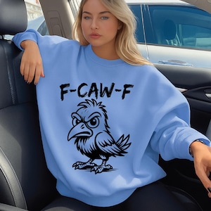 Puede incluir: Sudadera azul claro con el texto "F-CAW-F" y una ilustración negra de un pájaro enfadado. El pájaro tiene un gran pico y está de pie. La sudadera tiene mangas largas.