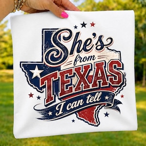 May include: A white t-shirt featuring a graphic of the state of Texas in red, white, and blue. The design includes the text She's from TEXAS I can tell in a stylized font, accented with stars and a distressed texture throughout the print.