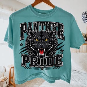 Op de afbeelding: Een t-shirt met korte mouwen in de kleur blauwgroen met een afbeelding van een zwarte panterkop met gele ogen en een open bek. De tekst PANTHER PRIDE staat in zwarte blokletters gedrukt, geaccentueerd door klauwsporen en sterren.