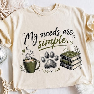 May include: A cream-colored t-shirt featuring the black and olive green text My needs are simple. The graphic includes a steaming olive green coffee mug, a black paw print, and a stack of three olive green books with decorative gold accents.