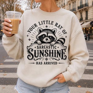 Puede incluir: Una sudadera beige de cuello redondo con un estampado gráfico negro de un mapache asomándose sobre un letrero. El texto dice: YOUR LITTLE RAY OF SARCASTIC SUNSHINE HAS ARRIVED. El diseño incluye estrellas decorativas y adornos alrededor del dibujo.