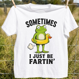 Può includere: Una t-shirt bianca a maniche corte con la grafica di una rana verde che indossa un salvagente giallo a forma di papera. Il testo SOMETIMES I JUST BE FARTIN' è presente con una piccola icona a forma di nuvola vicino al corpo della rana.