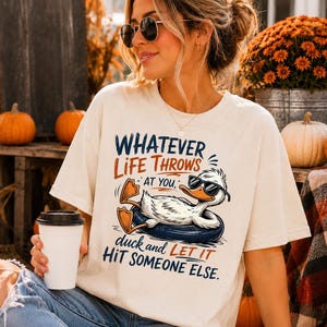 Woman wearing a cream oversized t-shirt featuring a funny duck in sunglasses relaxing on a float with the quote "Whatever Life Throws At You, Duck and Let It Hit Someone Else," styled with cozy autumn decor and pumpkins.
