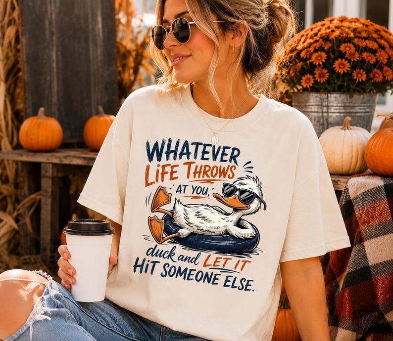 Woman wearing a cream oversized t-shirt featuring a funny duck in sunglasses relaxing on a float with the quote "Whatever Life Throws At You, Duck and Let It Hit Someone Else," styled with cozy autumn decor and pumpkins.
