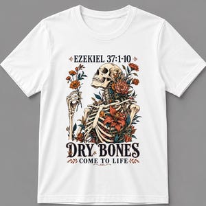 May include: A white short-sleeved t-shirt featuring a graphic of a human skeleton surrounded by orange and red flowers. The text EZEKIEL 37:1-10 appears above the skeleton, and the text DRY BONES COME TO LIFE is printed in black below the illustration.