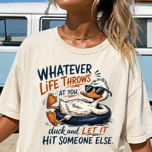 May include: A cream-colored t-shirt featuring a graphic of a white duck wearing black sunglasses while floating in a dark blue inner tube. The text reads: WHATEVER LIFE THROWS AT YOU, duck and LET IT HIT SOMEONE ELSE. The design uses blue and orange ink.