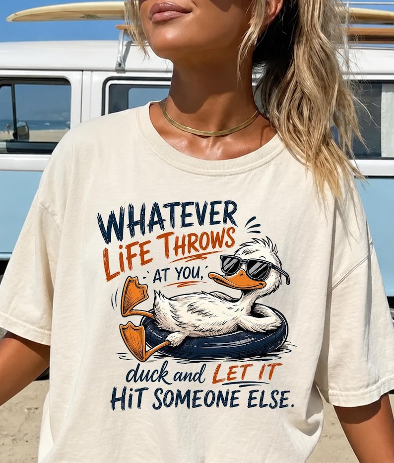 May include: A cream-colored t-shirt featuring a graphic of a white duck wearing black sunglasses while floating in a dark blue inner tube. The text reads: WHATEVER LIFE THROWS AT YOU, duck and LET IT HIT SOMEONE ELSE. The design uses blue and orange ink.