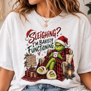 Op de afbeelding: Een wit t-shirt met een afbeelding van een groen wezen met een kerstmuts, zittend in een stoel onder een rood-zwart geruite deken. De tekst luidt: Sleighing? I'm Barely Functioning. Bevat een bordje met de tekst Hot Cocoa, Blankets, Maybe Later.