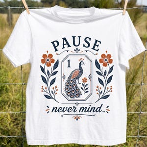 May include: A white short-sleeve t-shirt featuring a graphic print with the text PAUSE and never mind. The design includes a central peacock inside an octagonal frame with the number 1, flanked by orange flowers and dark blue leafy stems.