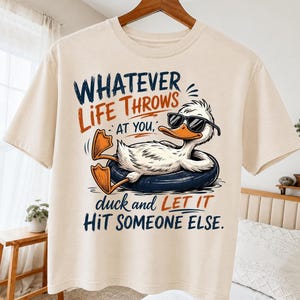 May include: A cream-colored t-shirt featuring a graphic of a white duck wearing black sunglasses and relaxing in a dark blue inner tube. The text reads: WHATEVER LIFE THROWS AT YOU, duck and LET IT HIT SOMEONE ELSE. The design uses navy and orange ink.