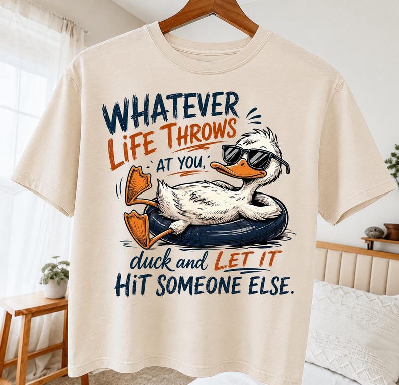 May include: A cream-colored t-shirt featuring a graphic of a white duck wearing black sunglasses and relaxing in a dark blue inner tube. The text reads: WHATEVER LIFE THROWS AT YOU, duck and LET IT HIT SOMEONE ELSE. The design uses navy and orange ink.