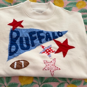 May include: A cream-coloured crewneck sweatshirt featuring a blue pennant flag with the word BUFFALO appliqued in dark blue fabric. The design includes three fabric stars in red, red-and-white gingham, and red floral print, plus a brown football.