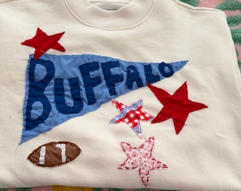 May include: A cream-colored crewneck sweatshirt featuring a blue pennant flag with the word BUFFALO appliqued in dark blue fabric. The design includes three fabric stars in red, red-and-white gingham, and red floral print, plus a brown football.