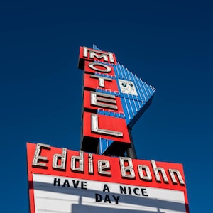 May include: A red and blue motel sign with white neon lettering that reads "Motel" and "Eddie Bohn". The sign also has a white message board that reads "Have a Nice Day".
