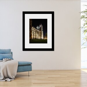 May include: A black and white photograph of a large, illuminated building at night. The building has a tall, pointed spire and many windows. The building is surrounded by a dark, grassy area.