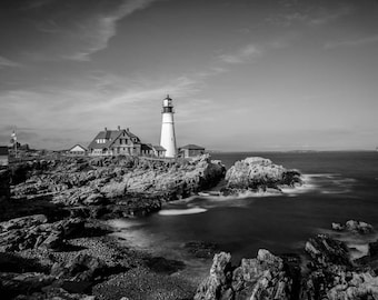 May include: A black and white photograph of a lighthouse on a rocky coast. The lighthouse is tall and white with a black roof. The water is choppy and the sky is cloudy.