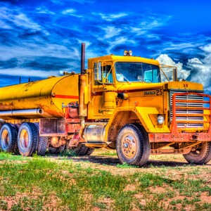 May include: A yellow tanker truck with a KW logo and the number 25 on the side. The truck is parked on a grassy field with a blue sky and white clouds in the background.