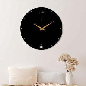 May include: A black, round wall clock with gold-colored hands and a minimalist design. The clock features the number 12 at the top and a small cutout at the bottom for the number 6. The clock is mounted on a white wall.