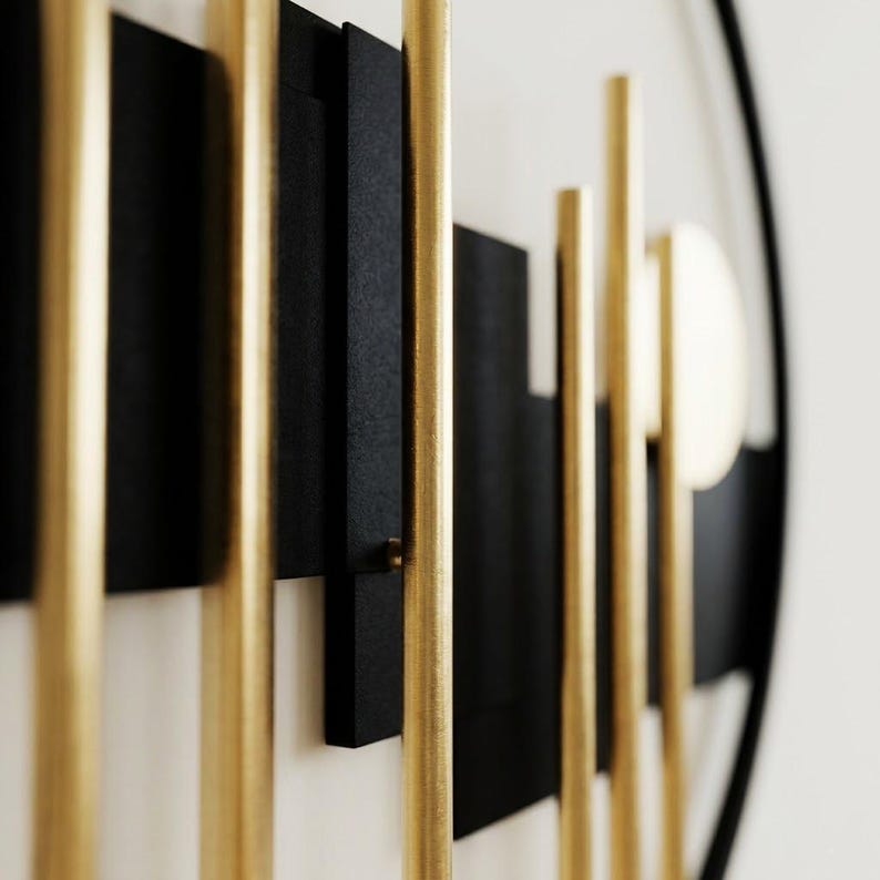 May include: A circular wall art piece featuring a black metal frame and a white background. The design includes vertical gold-toned metal rods of varying lengths layered over horizontal black rectangular panels and a circular gold-toned disc element.