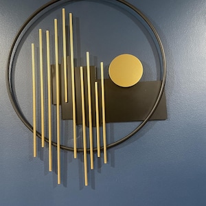 May include: A circular black metal wall art piece featuring a series of vertical gold-toned rods of varying lengths arranged in a linear pattern, accompanied by a solid black rectangular plate and a single gold-toned circular disc element.