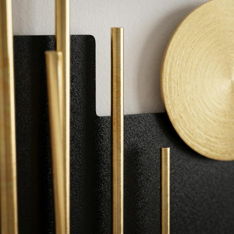 May include: A wall-mounted decorative piece featuring a textured black metal panel with vertical gold-toned cylindrical rods of varying heights and a large, circular gold-toned disc with a brushed finish positioned to the right side.