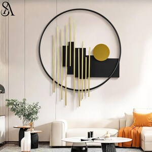 May include: A large circular wall art piece featuring a black metal frame, vertical gold-toned rods of varying lengths, black rectangular panels, and a solid gold-toned circle. The design is mounted on a wall above a white sofa and side table.