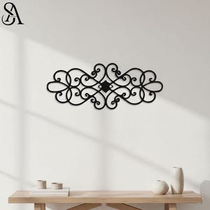 May include: Black metal wall decor with an ornate, symmetrical design. The piece features swirling patterns and a central diamond-shaped element. The decor is mounted on a white wall, with a wooden table and decorative vases in the foreground.