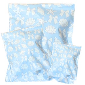 May include: A set of three light blue fabric bags with a repeating pattern of white seashells, starfish, and bows. The bags vary in size, with the largest bag in the background and the smallest in the foreground. The bags are likely for storage or gift wrapping.