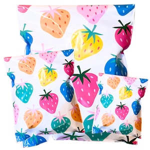 Puede incluir: Bolsas de embalaje blancas con un colorido estampado de fresas. Las fresas son de varios colores, incluyendo rosa, naranja, amarillo, azul y verde. Las bolsas son de diferentes tamaños y probablemente se utilizan para empaquetar regalos o productos.