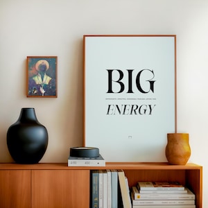以下が含まれることがあります： 黒い花瓶、小さな絵画、黒いボウル、そして「BIG ENERGY」と書かれた額入りのプリントがある木製のキャビネット。プリントは水色の背景と茶色のフレームです。本が棚に積まれています。