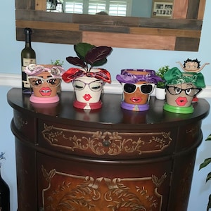 May include: Four painted terracotta flower pots with faces, sunglasses, and bandanas. The pots are arranged on a dark brown table with a carved design. One pot has a green bandana, one has a purple bandana, one has a red bandana, and one has a blue bandana.
