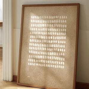 以下が含まれることがあります： ベージュ色の背景に、白の不規則な形が格子状に配置された、額装された抽象的なアートプリント。木製のフレームがアートワークのニュートラルな色調を引き立てています。壁に立てかけられており、ミニマリストなデザインが特徴です。