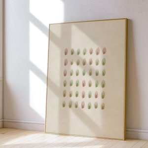 May include: A framed abstract art print featuring a pattern of small, colorful ovals in shades of green, pink, and brown. The artwork is set against a neutral background and framed in a gold-colored frame. The print is leaning against a white wall.