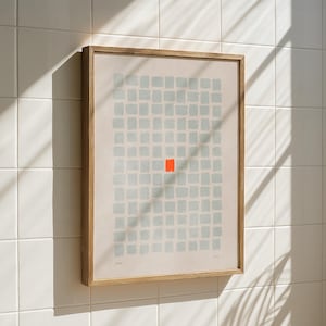 Könnte beinhalten: Abstrakter Kunstdruck in einem Holzrahmen, der ein Raster aus hellblauen Quadraten mit einem zentralen roten Quadrat zeigt. Das Kunstwerk ist vor einem beigen Hintergrund platziert. Der gerahmte Druck hängt an einer weiß gefliesten Wand.