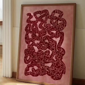 May include: Framed abstract art print. The artwork displays a complex pattern of dark red, intertwined shapes with white outlines, set against a soft pink background. The wooden frame leans against a wall, offering a modern aesthetic for home decor.