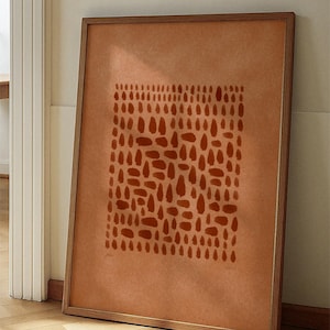 May include: A framed art print with an abstract design of rust-coloured shapes on a terracotta background. The wooden-framed artwork leans against a wall, offering a modern, minimalist aesthetic. The print is suitable for home decor.
