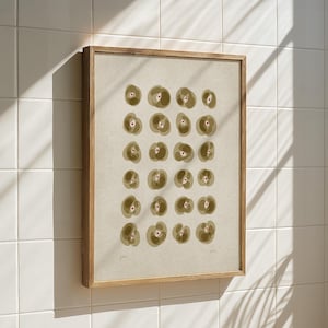 May include: A framed art print featuring a beige background with a pattern of olive-green and pink floral-like shapes. The artwork is encased in a light brown wooden frame and hangs on a white tiled wall.