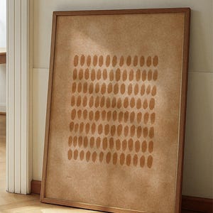 Può includere: Una stampa artistica astratta incorniciata con un motivo di forme ovali marroni su sfondo beige. L'opera è esposta in una cornice di legno e appoggiata a un muro, mostrando un design minimalista.
