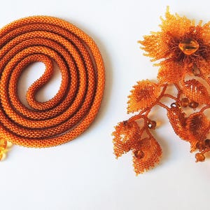 May include: An orange beaded necklace coiled in a spiral next to an orange beaded floral brooch. The necklace has a beaded fringe with orange flower-shaped beads. The brooch features a large flower and leaf design.