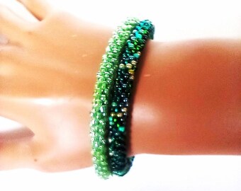 May include: A green beaded bracelet, with varying shades from light to dark, encircles a wrist. The bracelet is made of small, round beads, giving it a textured appearance. The beads catch the light, creating a subtle sparkle in the design.